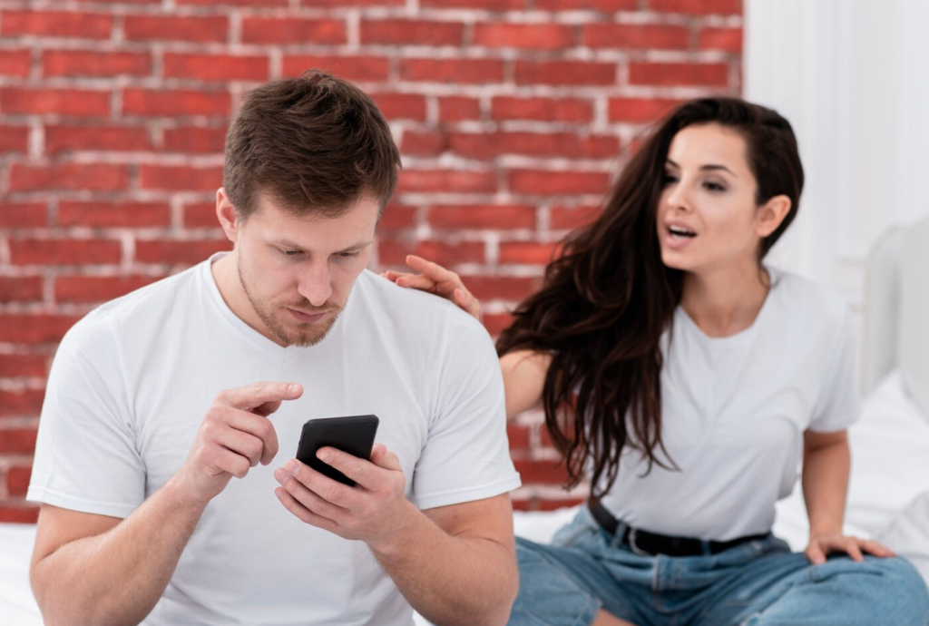 Man focused on smartphone while woman gestures towards him, sitting on bed with a red brick wall background.