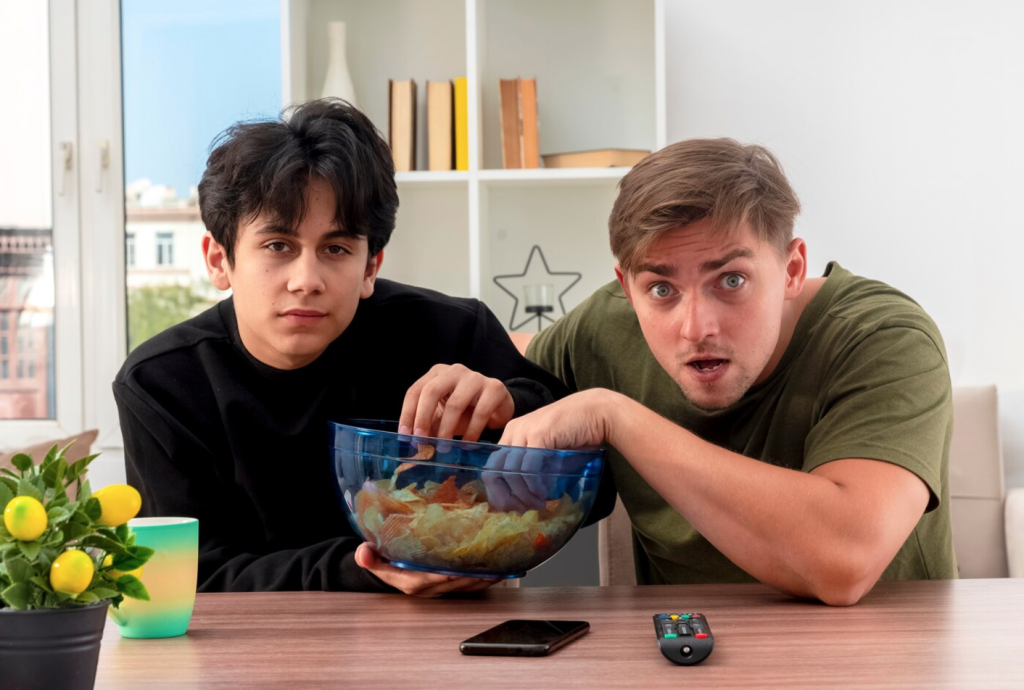 Two men sitting at a table with a bowl of chips, one looking surprised. Remote and phone on the table.