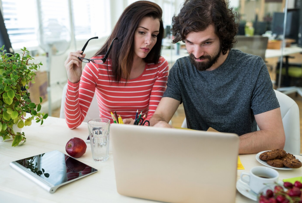 Man and woman collaborating at a laptop, surrounded by plants, snacks, and stationery on a bright office desk.