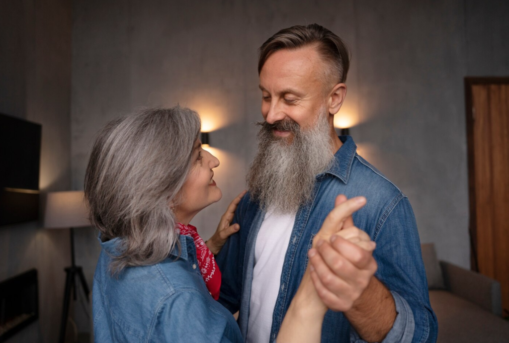Older couple in denim shirts joyfully dancing together in a cozy, warmly lit room.