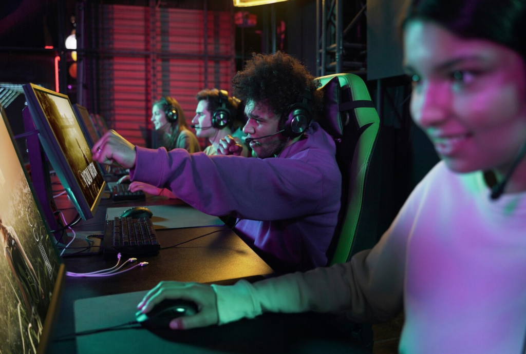 Gamers wearing headsets playing on computers in a dimly lit room with neon lighting.