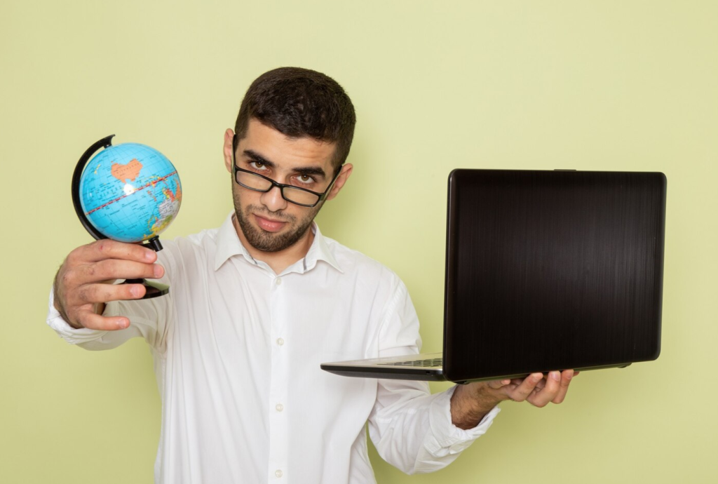 Man holding a small globe and a laptop against a light green background, wearing glasses and a white shirt.