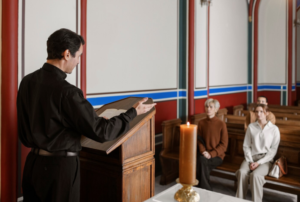 A person reads from a lectern in a church, with a candle beside them, addressing a small seated congregation.