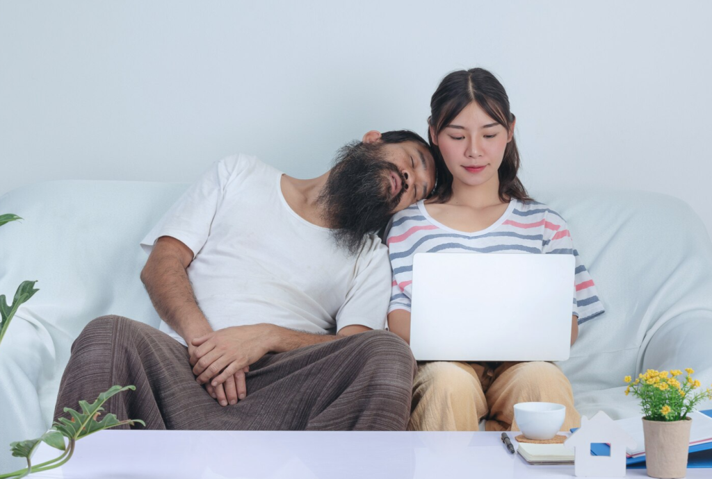 Woman uses laptop on couch, while man leans asleep on her shoulder; plant and mug on table nearby.