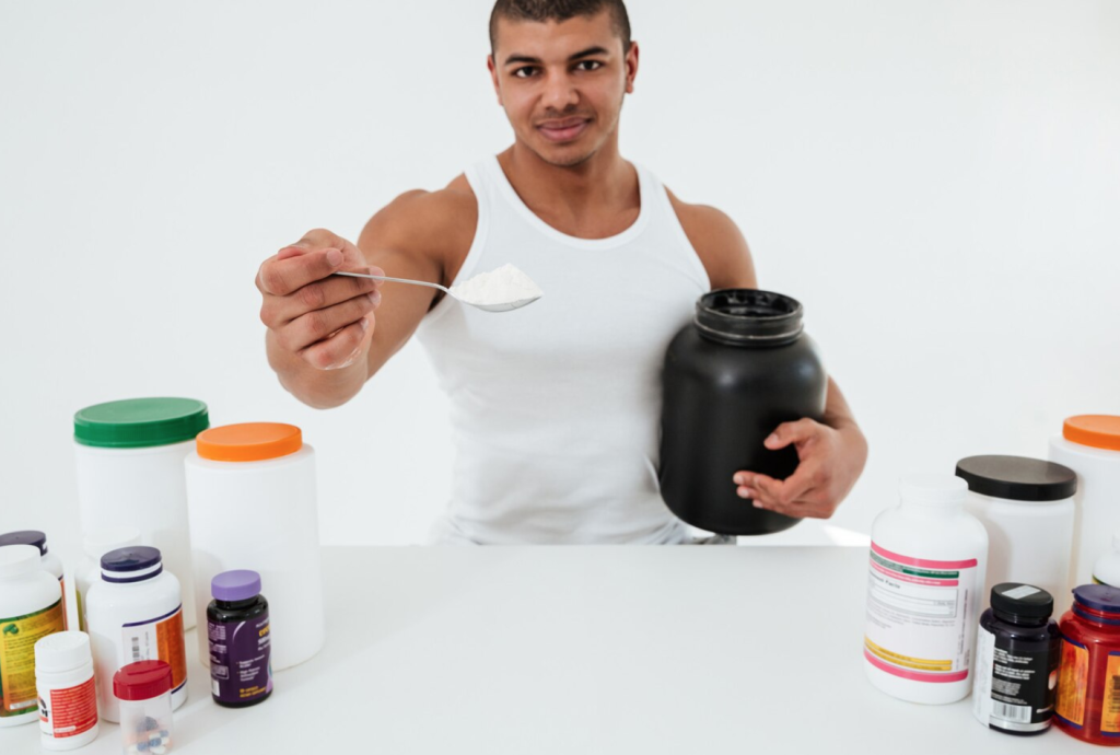 Man in white tank top holding spoon of powder with supplement containers on a table.