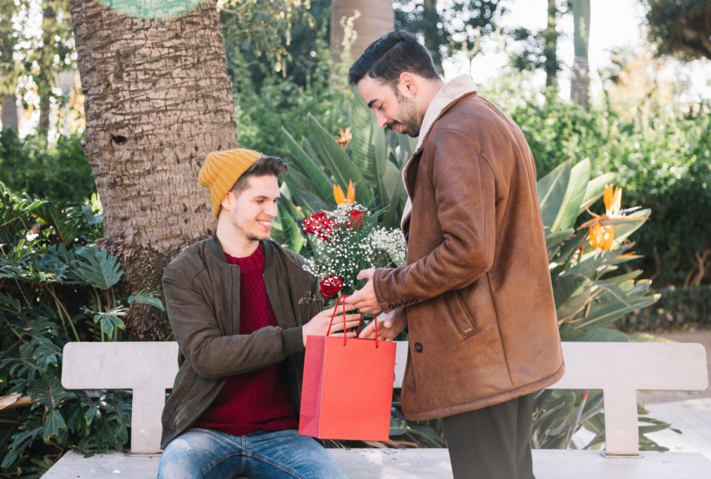 Two men outdoors; one gives the other flowers and a gift bag while sitting on a bench surrounded by greenery.