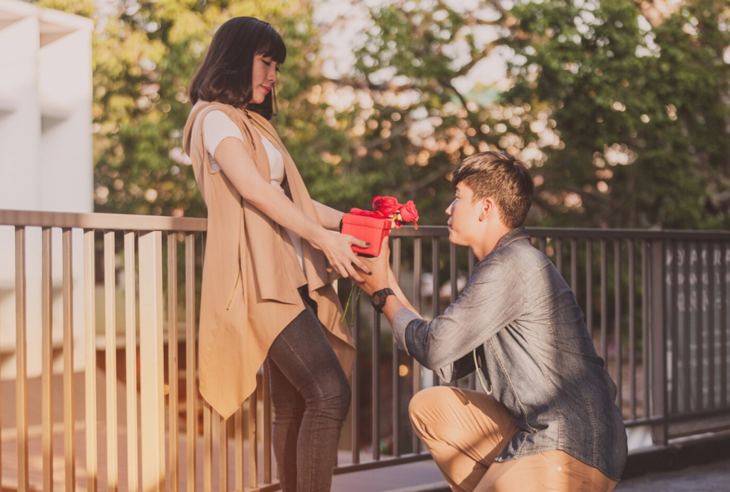 Man kneeling offers red roses to woman on a bridge during sunset.