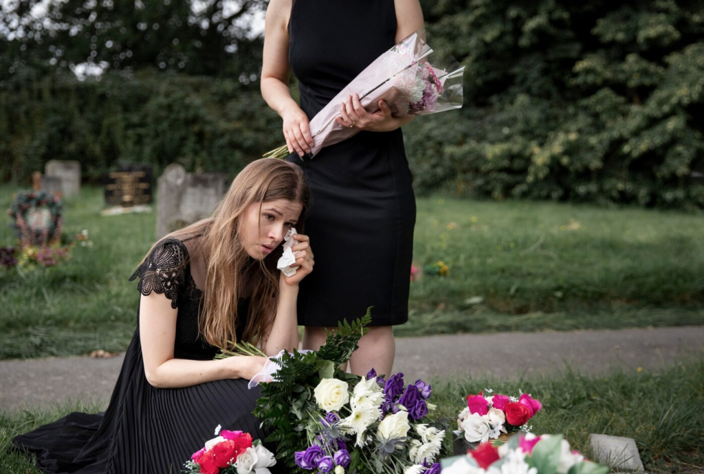 Two women in black mourn at a gravesite, one kneeling with a tissue and flowers, the other standing with a bouquet.