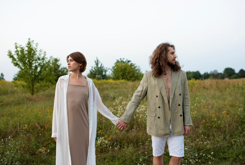 A couple holding hands in a grassy field, both looking away, wearing neutral-toned outfits.