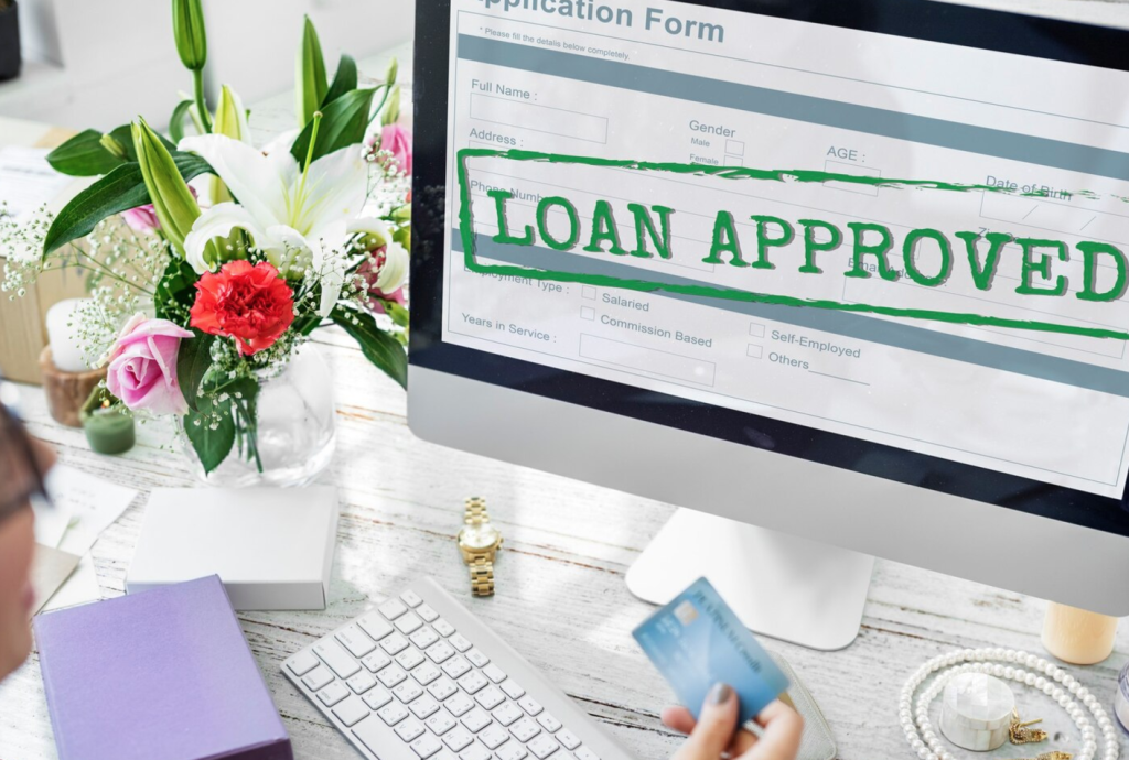Computer screen displaying "Loan Approved" with a credit card in hand and flowers on the desk.