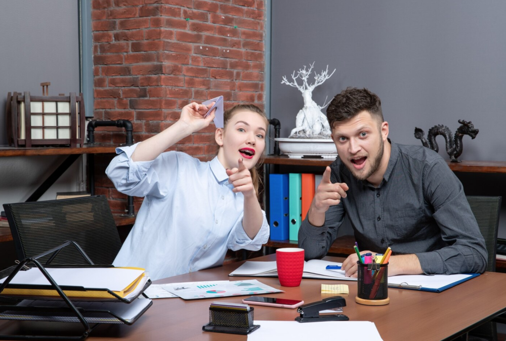 Two colleagues at a desk, smiling and pointing playfully, with office supplies and a brick wall in the background.