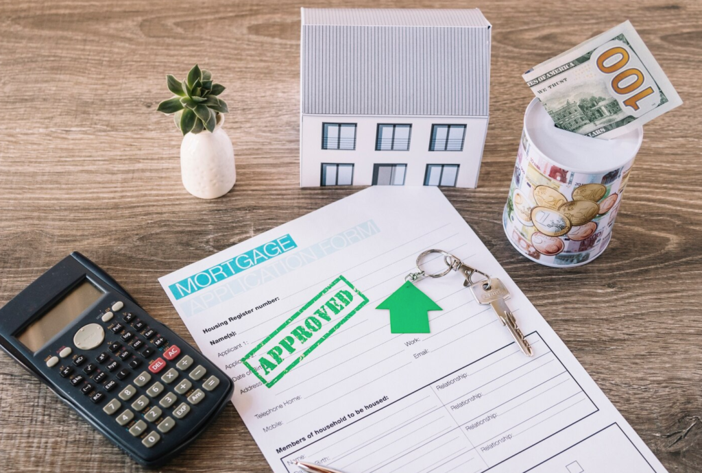Mortgage application marked "Approved" with keys, calculator, model house, and a money jar on a wooden table.