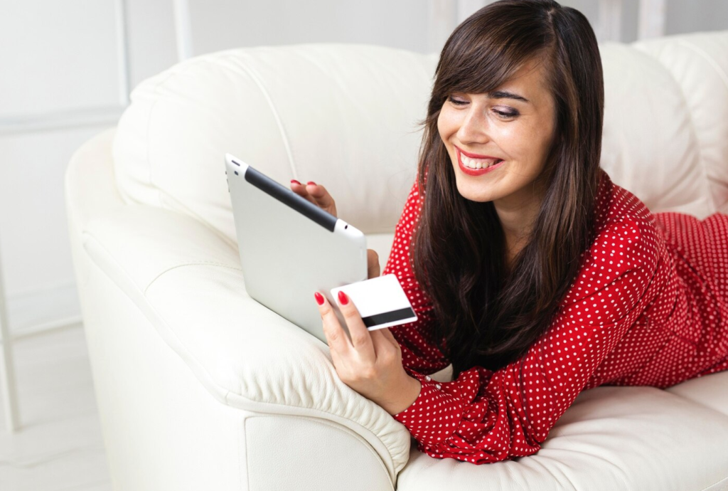 Woman in red polka dot dress shopping online with a tablet and credit card, smiling on a white couch.
