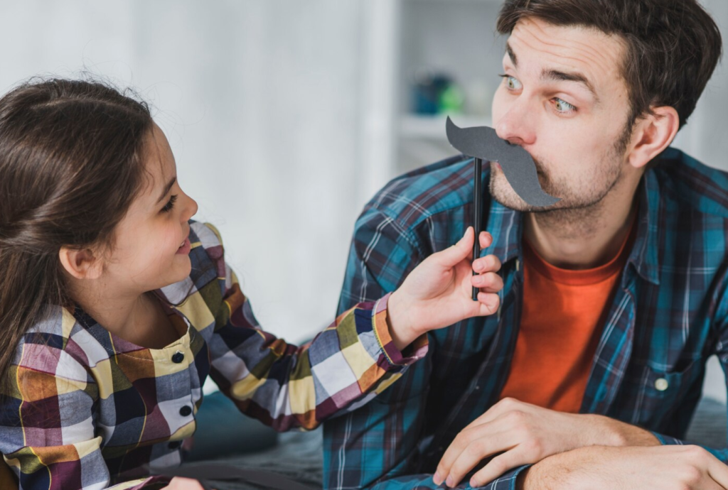 Child playfully holds a fake mustache to a man's face, both wearing plaid shirts and smiling at each other.