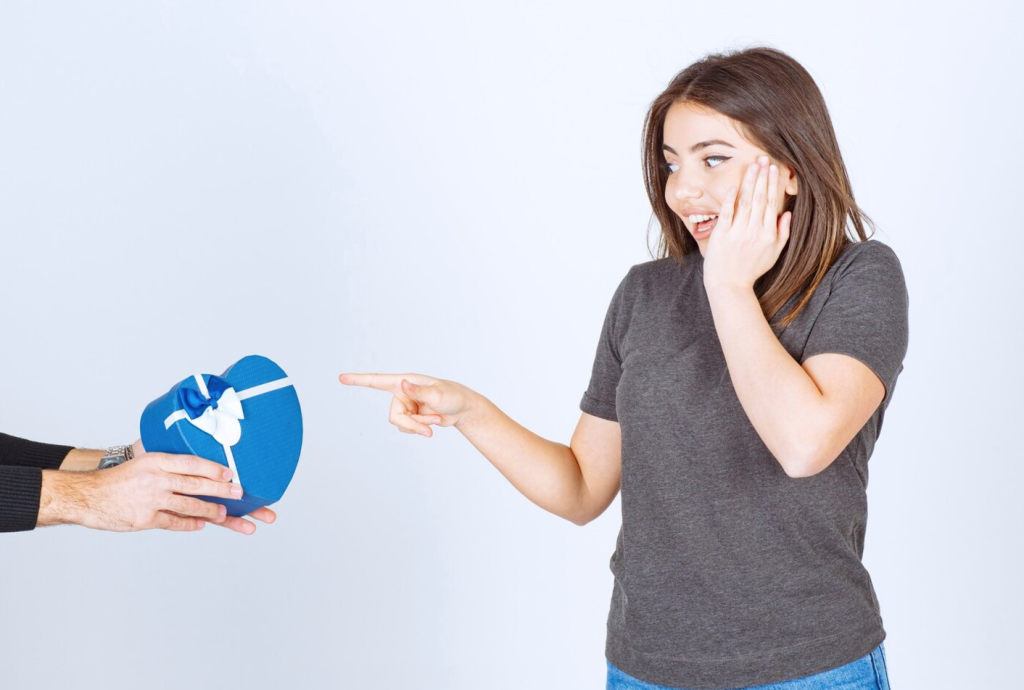 Surprised woman pointing at a blue heart-shaped gift box being offered by another person.