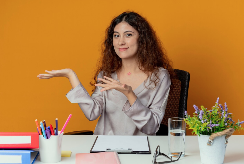 Woman with curly hair sits at a desk with office supplies, gesturing with hands, in front of an orange background.