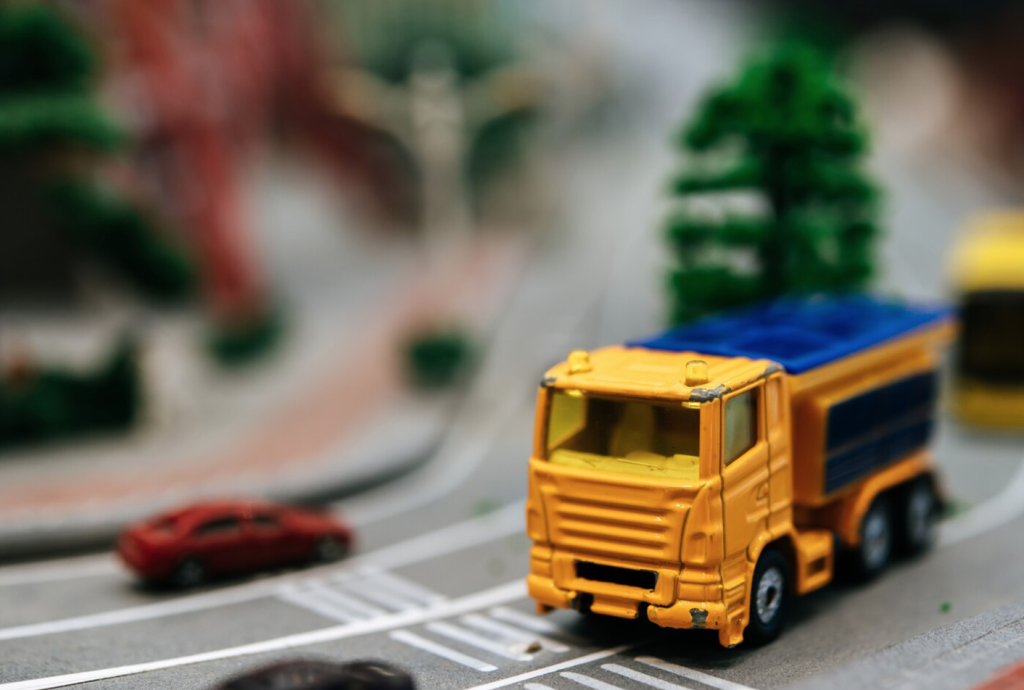 Toy yellow truck on a miniature road with blurred background featuring trees and a parked red car.