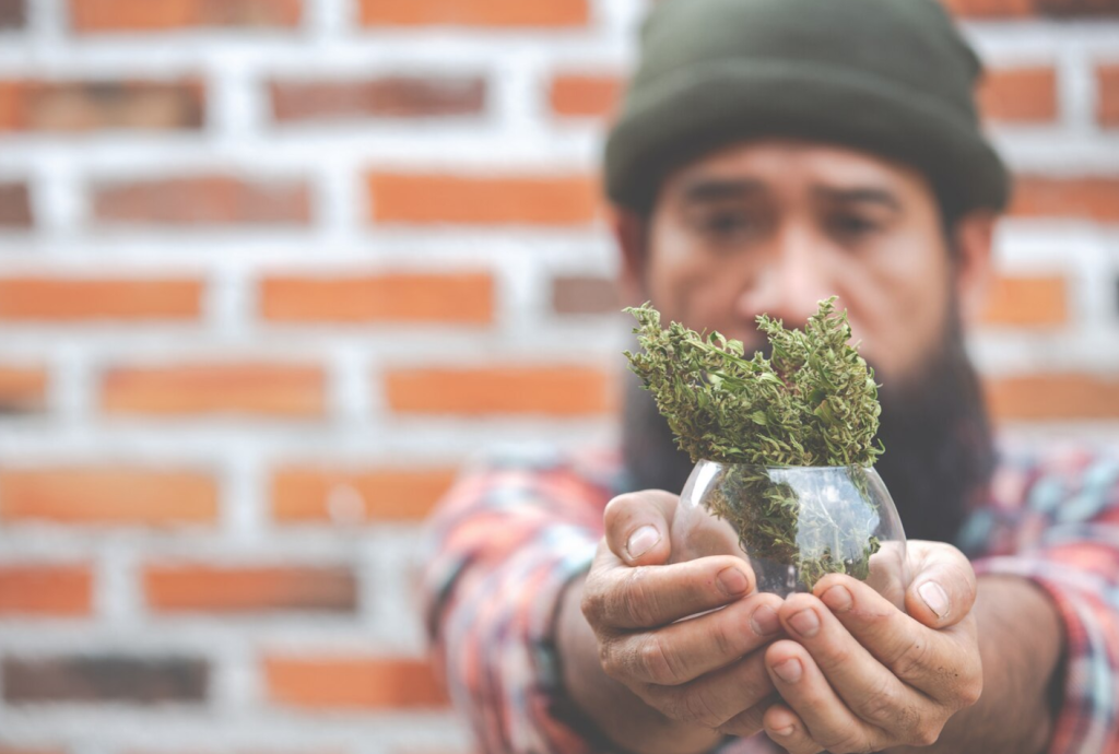 Person holding a small glass vase with dried herbs, standing in front of a brick wall.