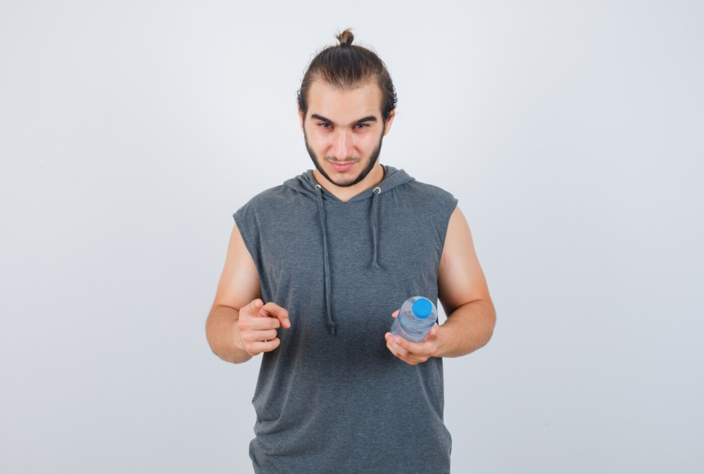 Man in gray sleeveless hoodie holding a water bottle and pointing, standing against a plain background.