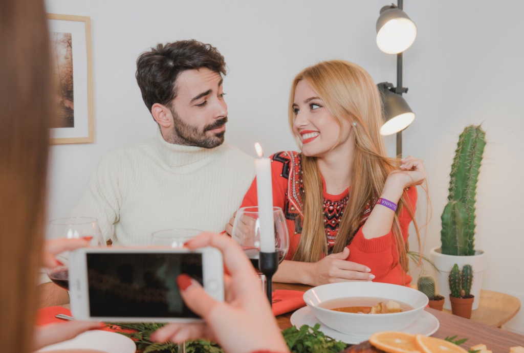 A couple smiles at each other during dinner, while someone takes a photo with a smartphone.