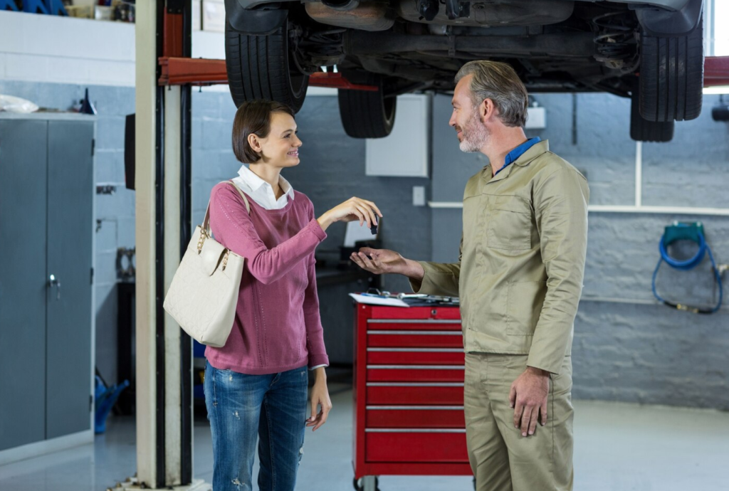 Woman giving car keys to a mechanic in a garage; car lift and red tool chest in the background.