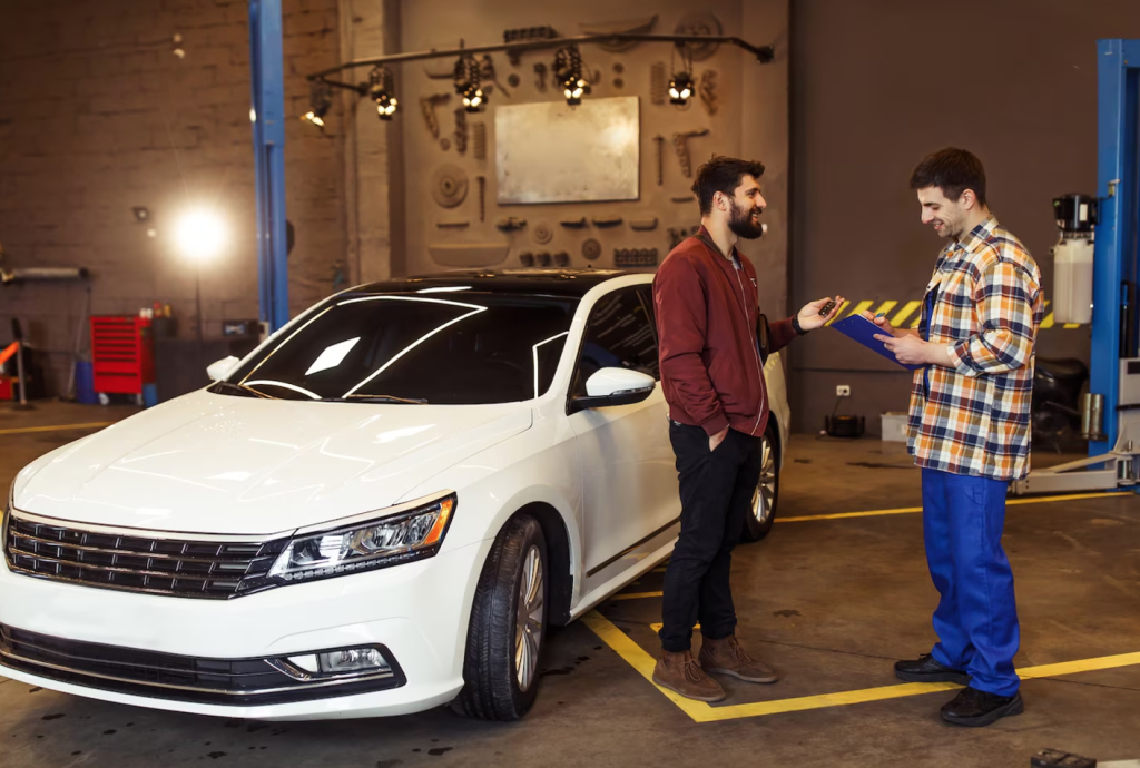 Two men talking beside a white car in an auto repair shop, with one holding a clipboard.