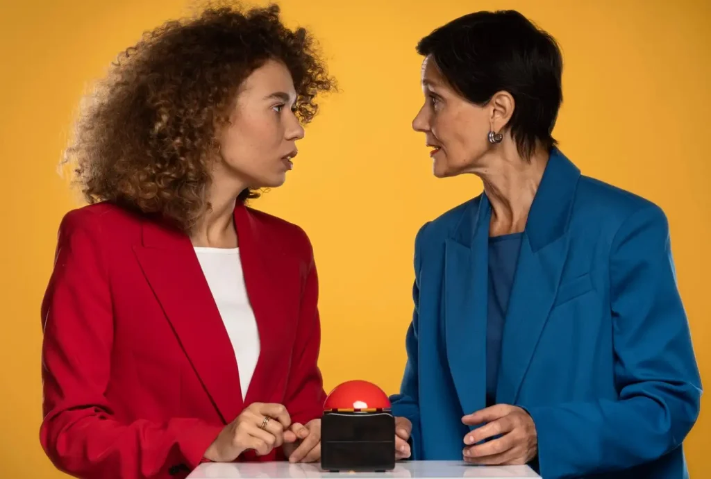 Two women in suits facing each other over a red button, set against a yellow background.
