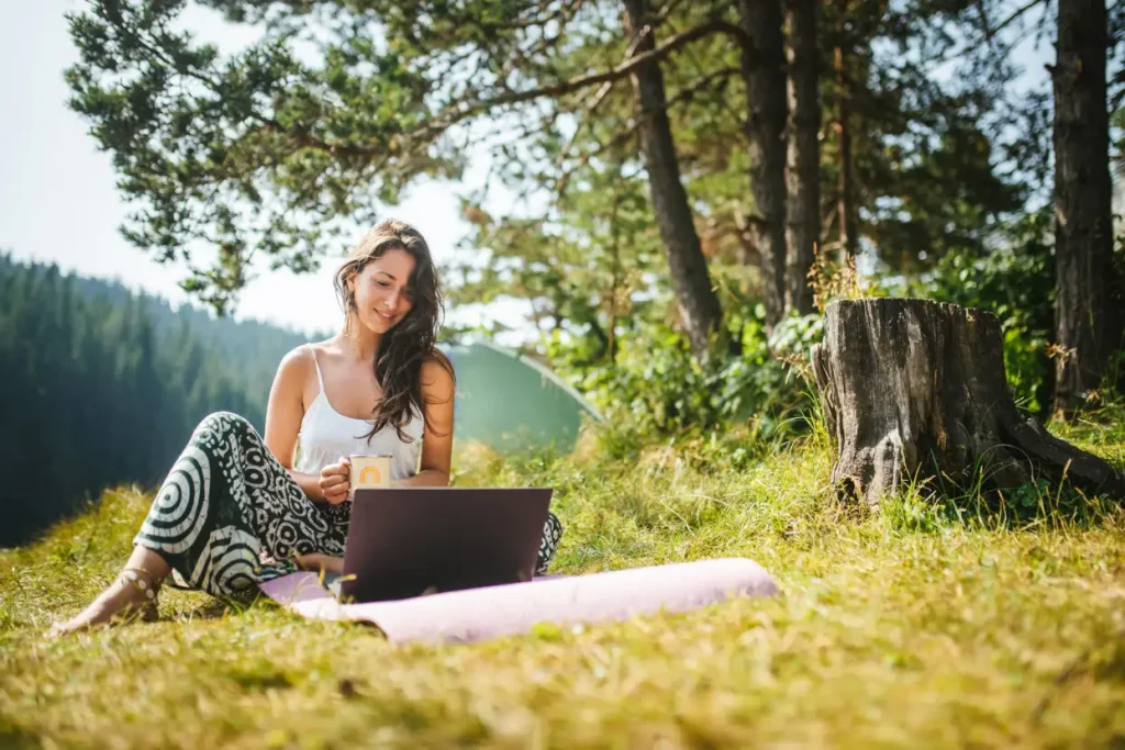 Woman sitting on grass with a laptop and mug, surrounded by trees, enjoying a sunny day outdoors.