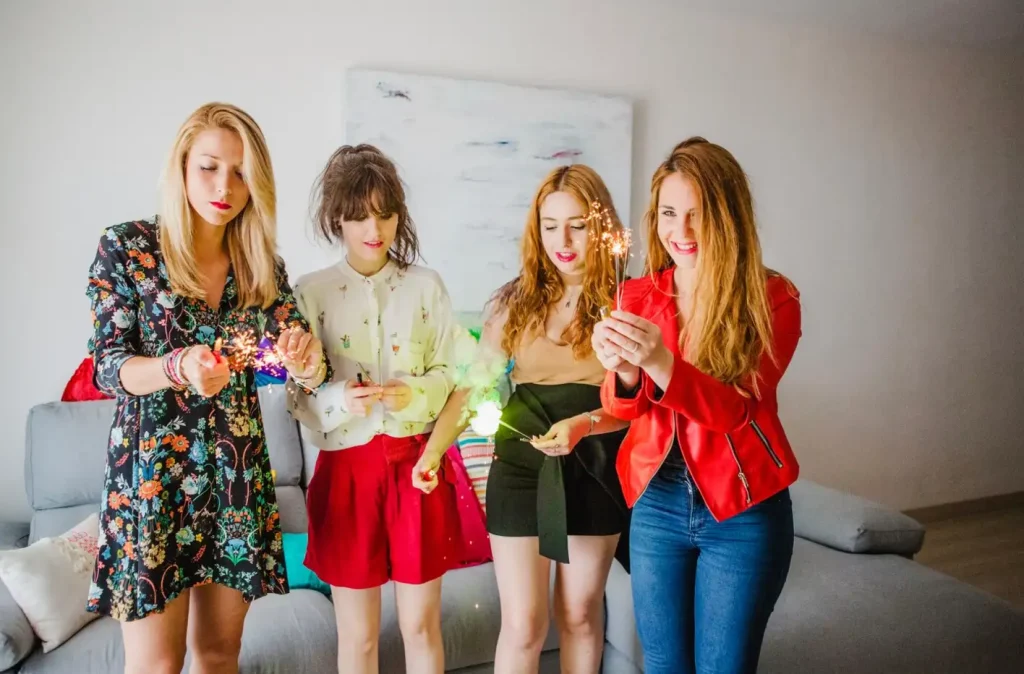 Four women indoors holding sparklers, smiling, and celebrating. The room has a sofa and abstract wall art in the background.