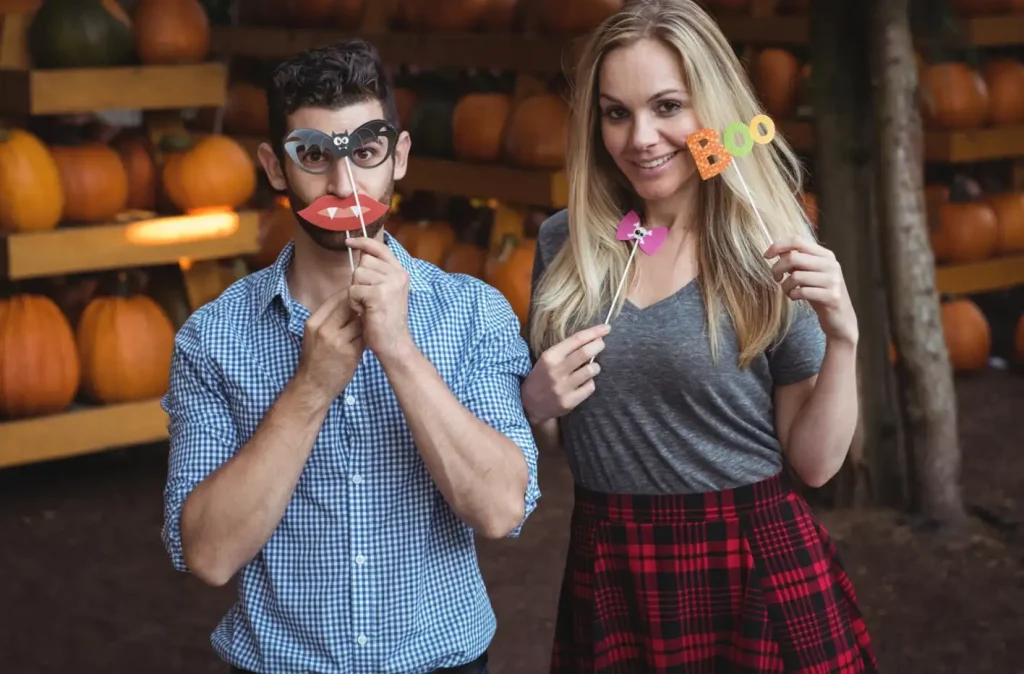 Two people at a pumpkin patch holding Halloween-themed props with playful expressions.