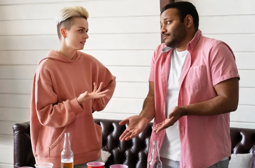 Two people gesturing while having a conversation at a table with bottles and cups in a casual setting.