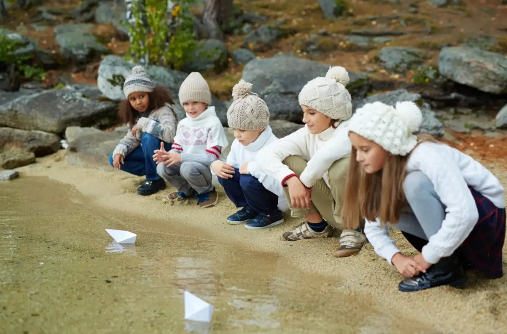 Children wearing knit hats kneeling by a lake, playing with paper boats on a sandy shore.