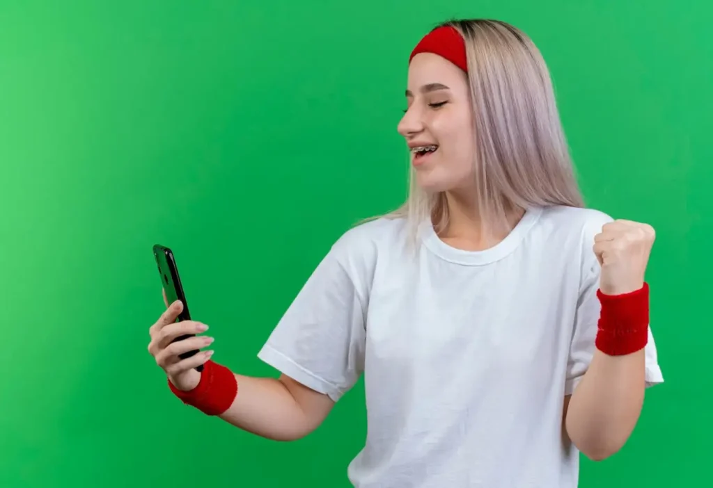 A person with a red headband and wristbands is joyfully looking at a smartphone, set against a green background.