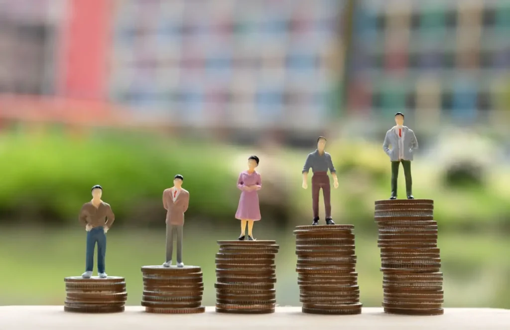 Miniature figures stand on stacks of coins, depicting various heights, symbolizing financial growth or economic disparity.