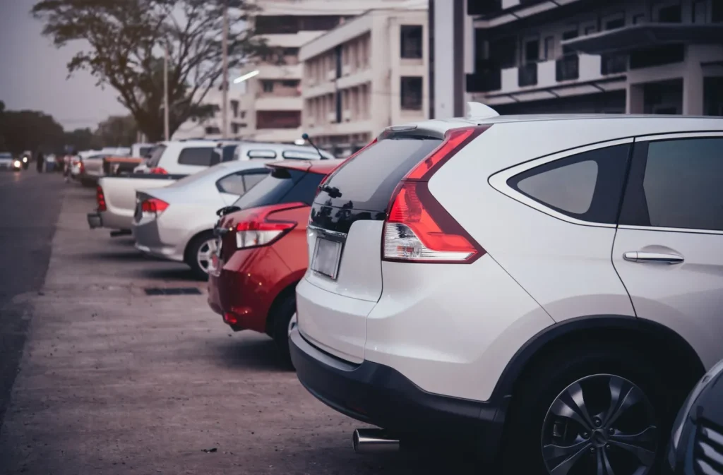 Row of parked cars in a city lot with buildings and trees in the background.