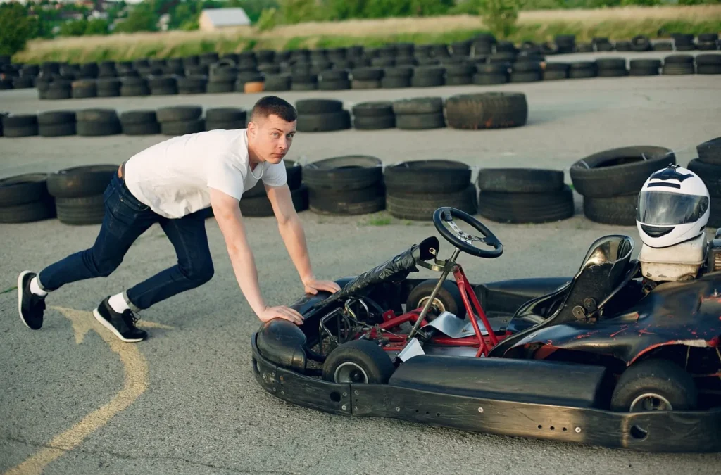 Man in a white shirt pushes a go-kart on a track surrounded by stacked tires under a clear sky.