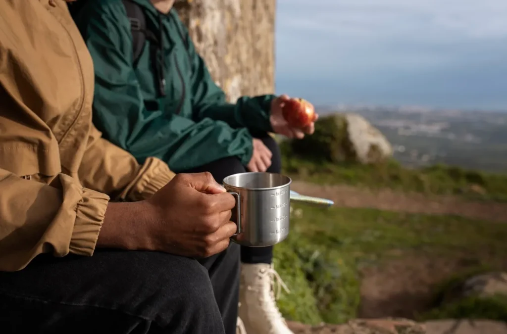 Two people sitting outdoors, one holding a metal mug and the other an apple, with a scenic landscape in the background.