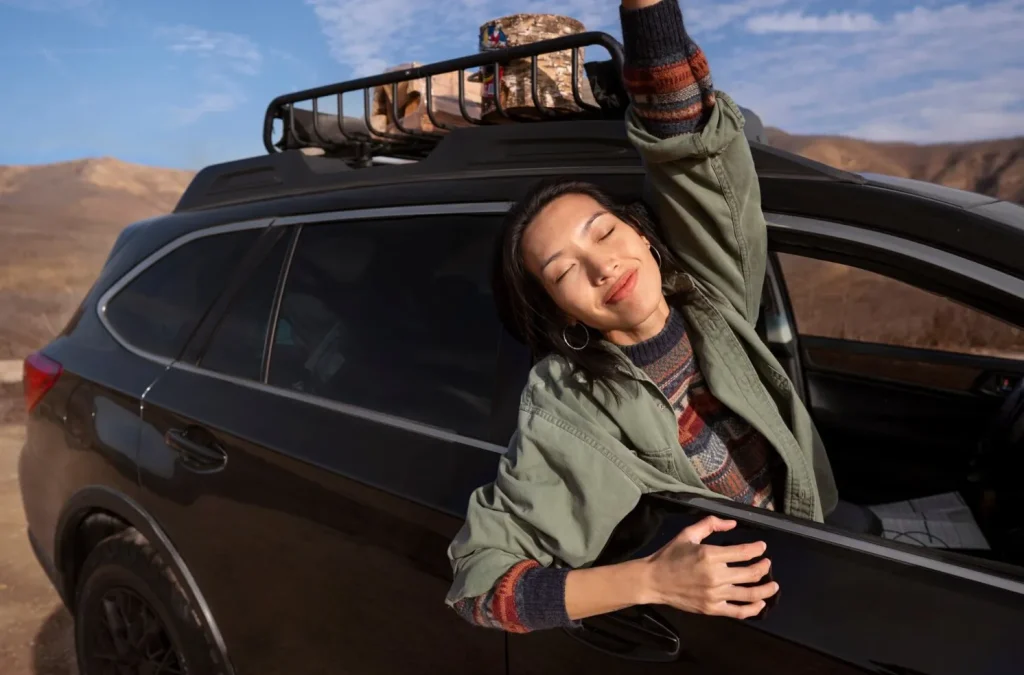 Woman smiling with eyes closed, leaning out of a car window on a sunny day, with mountains in the background.