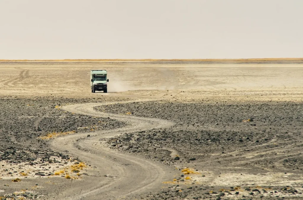 Truck driving on a dusty, winding dirt road in a barren desert landscape under a clear sky.