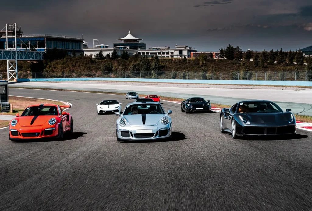 A group of sports cars speeding on a racetrack under a cloudy sky, with buildings and trees in the background.