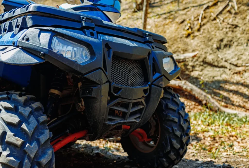 Close-up of an all-terrain vehicle's front, showing rugged tires and grille, on a sunny day in a forest setting.
