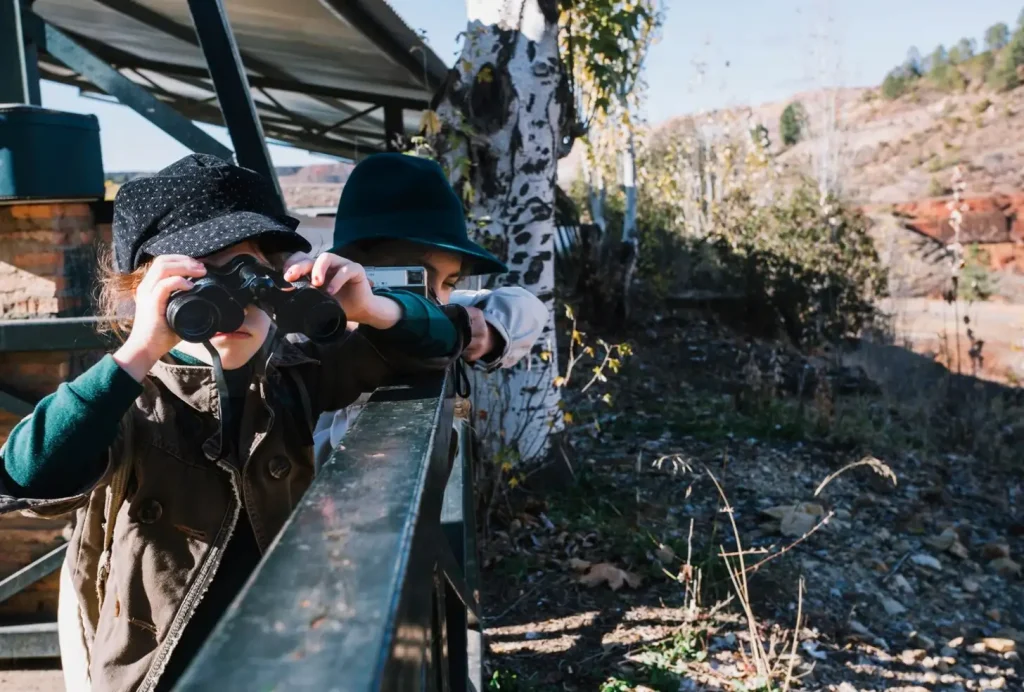 Two children with hats use binoculars and a camera to explore outdoors on a sunny day near trees and a building.
