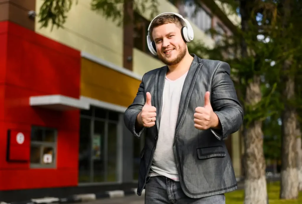 Man in casual suit and headphones gives thumbs up outside a colorful building.