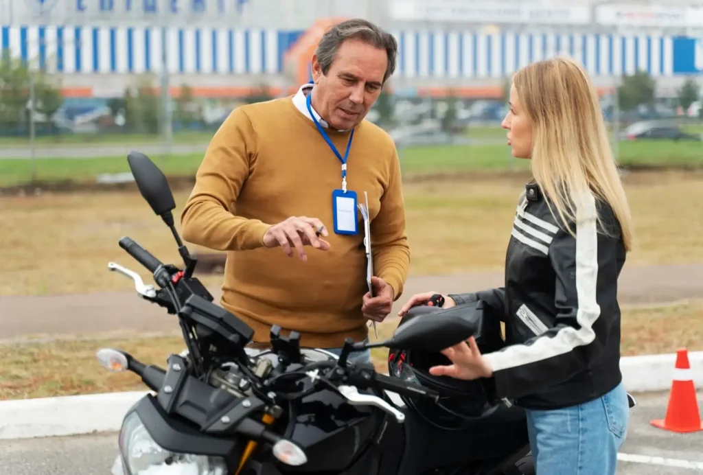 Man instructing woman on motorcycle handling outdoors, with buildings and traffic cones in the background.
