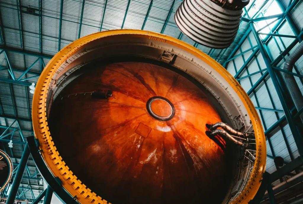 Large rocket fuel tank interior with orange tones, situated in an industrial hangar with a visible ceiling structure.