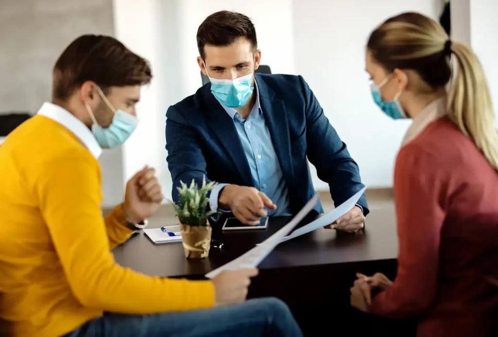 Three people in masks discuss documents around a table, with a small plant and notepad visible in the foreground.