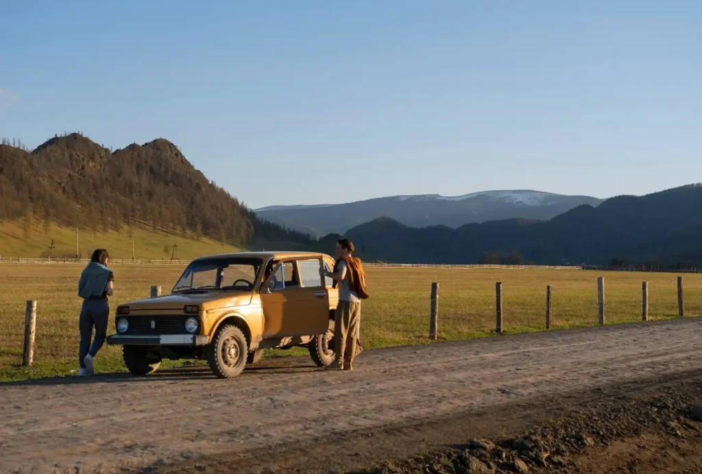 Two people stand by an orange car on a rural road, with mountains and fields in the background.