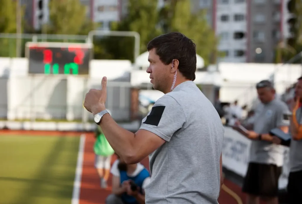 Coach giving a thumbs-up, back to a sports field, with a scoreboard showing 3-2 in the background.