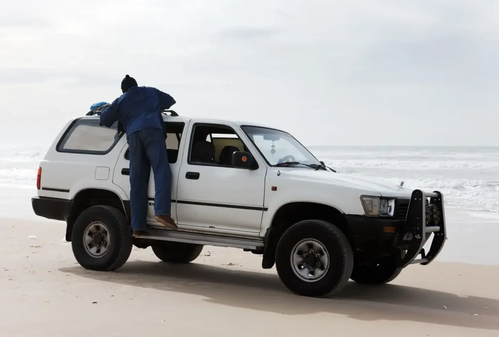 Person leans into white SUV parked on a sandy beach with ocean waves in the background.
