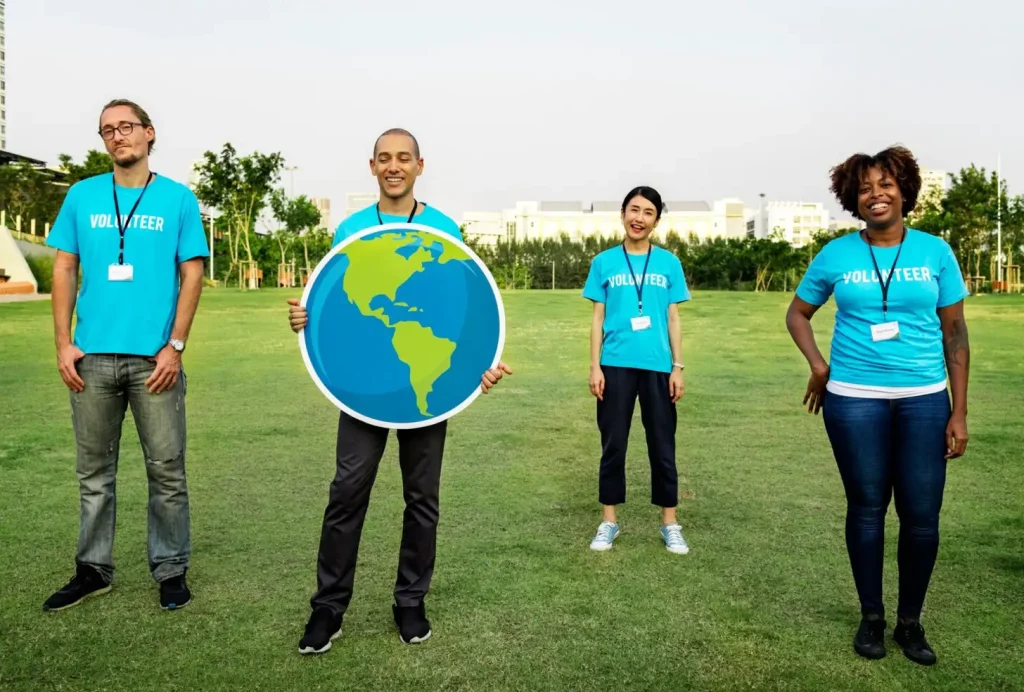 Four volunteers in blue shirts stand on grass, holding a globe cutout, with trees and buildings in the background.